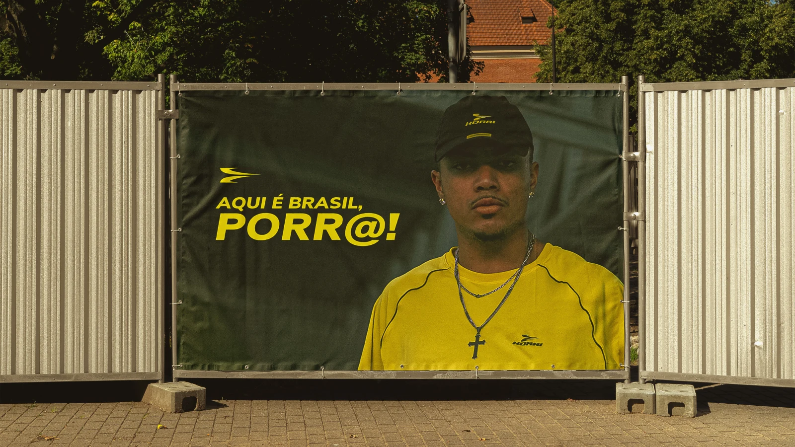 Banner em tapume com a frase "AQUI É BRASIL, PORR@!" ao lado de um modelo de camisa amarela.